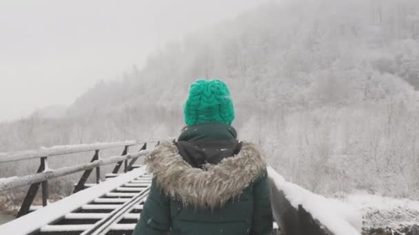 若い女性が雪の鉄道コースを散策し 周辺の自然と冬の休日を楽しんでいます ビデオは冬の散歩の美しさをキャプチャし 女性は雪に覆われた橋で楽しく旅行します — ストック動画