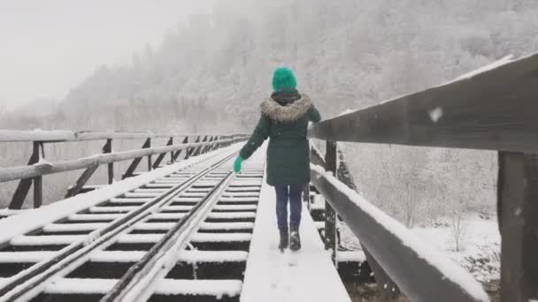 一个被雪覆盖的铁路桥 一个年轻的女人在被雪覆盖的树木间漫步 给观众一种和平与美丽的自然景观 一个心情愉快的姑娘沿着冬天的铁路桥走着 — 图库视频影像