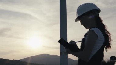Bir kadın mühendis, alternatif bir enerji santralindeki rüzgar türbininin enerji performansını analiz etmek ve veri toplamak için modern teknolojiyi kullanır. Güneşin doğuşunda siluet, kasklı adam. Yüksek kalite 4k