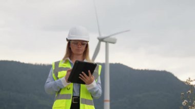 Yeşil enerji için dijital çözümler: bir tablet üzerindeki türbin ve yel değirmenlerinin analizi. Rüzgar enerjisi: Rüzgâr türbinleri, rüzgar türbini çiftliğinde yürüyen bir kadın bilim adamı tarafından yönetiliyor. Yüksek kalite 4k