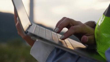 Yel değirmeni yakınında dizüstü bilgisayarı olan bir kadın: rüzgar çiftliği kurmanın teknik yönleri. Dizüstü bilgisayarın klavyesine bilgi yazıp elinizde tutuyorsunuz. Çevrimiçi veri izleme, online istatistik analizi. Yüksek