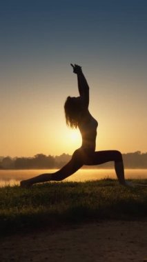 Güneşin doğuşundaki dikey videoda ince kadın silueti. Enerjiyi ve canlılığı arttırmak için yoga yapıyorum. Zihin ve beden uyum içinde: göleti bölen açık hava yogası. Nefes kesici güzel vücut figürü.