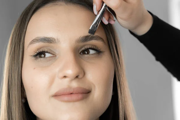 The photo captures the moment when the makeup artist works on every detail, creating eyebrows that emphasize the individuality and natural beauty of the face of a beautifully woman in a beauty salon
