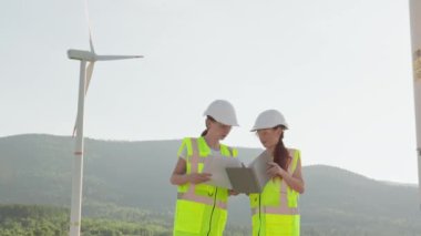 İnsanların bir yel değirmeninin, bir elektrik jeneratörünün arka planında durduğu genel bir plan. Çalışanlar, özel giysili mühendisler, miğferler ve yelekler teknik teftiş ve değerlendirme yaparlar.