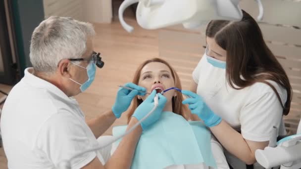 歯科医とアシスタントは歯の治療に慎重に取り組んでおり 患者の快適さを確保しています 歯科処置は診療所の現代内部で行われ 患者は感じます — ストック動画