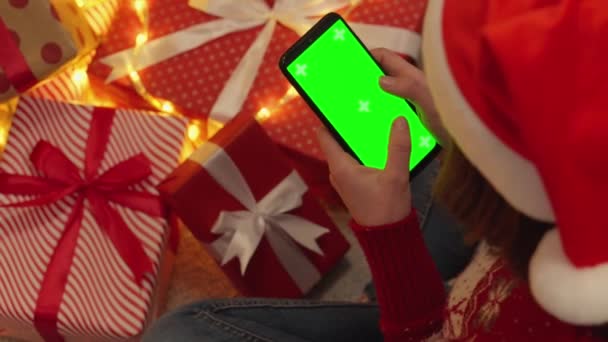 在圣诞树下 在礼品盒附近 一个戴着圣诞老人帽的女人和假日毛衣商店在一家网上商店里 用她的智能手机浏览促销活动 Chromakey效果和绿色屏幕 — 图库视频影像