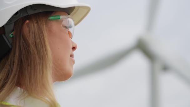 風力タービンはエネルギーの未来です ヘルメットとゴーグルに乗っている女性は 再生可能エネルギー発電所の発電機を慎重に調べます 持続可能な未来のための風力タービン エンジニアが座標 — ストック動画