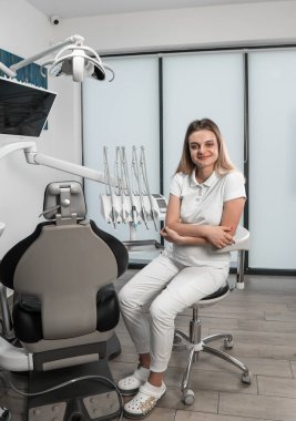 Bir dişçi, dişçi koltuğunun arka planına karşı durarak profesyonelliği ve kendine güveni gösterir. Bir dişçi modern teknolojileri kullanarak dişlerini düzeltir. Yüksek kalite fotoğraf