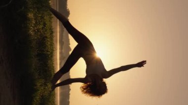 Kadın, doğada yoga sırasında kaslarını esnetiyor ve dengeyi canlandırıyor. Sağlık için Beden Eğitimi, Yaz Yogası Uygulaması: Bir kadın doğanın seslerini dinlerken asana yapar. Dikey