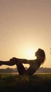 Gün doğumunda göl kenarında taze sabah havası soluyan kadın, vücut ve ruhun uyum ve rahatlaması için yoga yapıyor ve yapıyor. Asana çalan bir kadının dikey videosu.