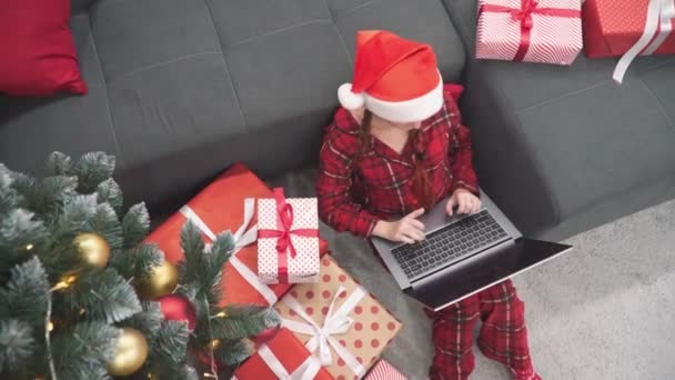 ガーランドやギフトで覆われたクリスマスウィガードの木の環境では 女性はラップトップで作業する快適さを楽しみ 重要な購入を行い 休暇を計画しています 高品質4Kについて — ストック動画