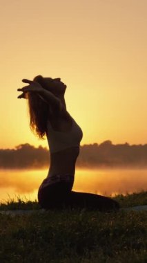 Doğa fiziksel ve ruhsal gelişim için bir ilham kaynağıdır. Yoga pratiği ve ince kadının dikey video siluetinde meditasyon için arka plan olarak gölün üzerinde gün batımı aydınlığı.