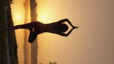 Dinlenme ve Konsantrasyon: Modern Bir Kadının Yaşamında Yoga Ritüeli. Rahatlamak ve formda kalmak için en iyi yol açık hava yogasıdır. Güneşin doğuşundaki seksi kadın figürünün dikey videosu. Yüksek kalite