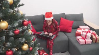 Çelenkler ve hediyelerle süslenmiş bir Noel ağacının sihirli ışığı altında, bir kadın dizüstü bilgisayarıyla çalışarak ve çevrimiçi bir bağlantı sağlayarak Noel havasını kutlamaya yardımcı oluyor. Yüksek kalite 4k