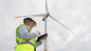 Kadın bir mühendis elindeki tabletle yenilenebilir enerjinin anahtar sürücüsü olarak rüzgarı keşfediyor. Yeşil bir gelecek için dijital araçlar: elektrik üretmek için kullanılan tablet ve yel değirmeni. Yüksek kalite 4k görüntü