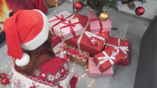 クリスマスのエレガンスをパーソナライズする女性は お祝いの精神を共有する準備ができて 赤いリボンでスタイリッシュにカーブされた贈り物を保持しています 女の子はクリスマスツリーの下に贈り物をします 高品質の4K映像 — ストック動画