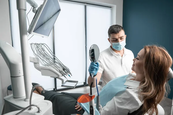 Doktor aynayı hastaya, kadına gösterir. Diş sağlığı merkezindeki prosedürler maksimum sonucu elde etmek için detaylara dikkat ve rahatlıkla uygulanır. Yüksek kalite fotoğraf