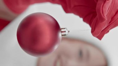 Dikey video. Noel ağacı süslemesi, elinde kırmızı eldivenli bir kızın elinde kırmızı bir balon. Önündeki kamerayı gösteriyor ve gülümsüyor. Noel ve Yeni Yıl kavramı