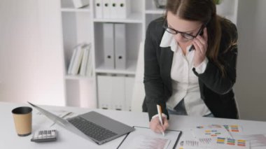Successful businesswoman in finance office working diligently on project analysis and planning. Businesswoman talking on mobile phone while standing at table. A woman fills out documents with a pen