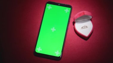 On a red background is a smartphone with a green screen with a wedding ring in a jewelry box. Valentines Day proposal and new year gifts to propose marriage to your loved one. High quality 4k footage