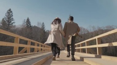 Bahar, aşk ve güneş. Romantik bir yürüyüş. Ilık ışıkla aydınlatılmış, birlikte vakit geçirmekten hoşlanan bir yürüyüş. Genç bir çift ahşap bir köprüde neşeyle koşuyor. Doğada eğlenmekten mutluyum. Yüksek kalite 4k