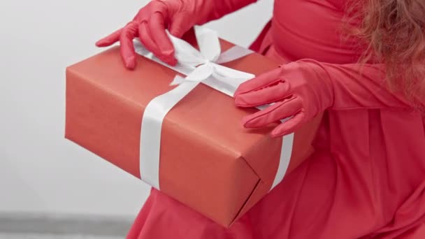 赤い夜の服を着た女性が誕生日プレゼントを受け取りました 彼は包装されたパッケージの白いリボンの弓を手で調整する 高品質の4K映像 — ストック動画