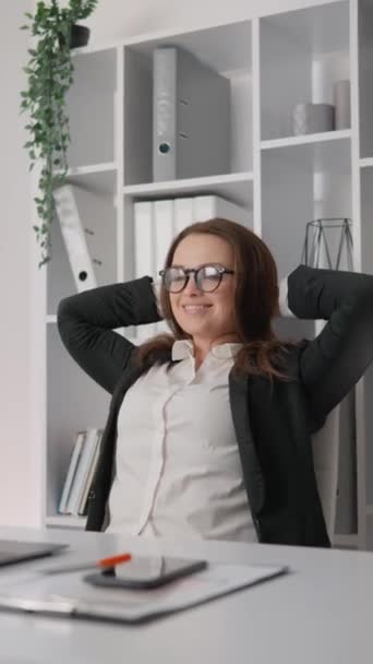 職場の職場で机に座りながら笑顔のプロのビジネスマン 女性は喜んでストレッチし ビジネスススーツでオフィスの椅子に座っています 労働日の終わり 成功しました — ストック動画