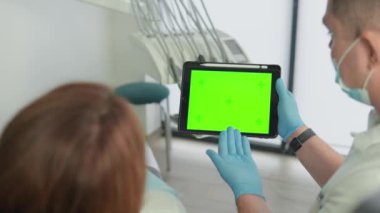 Diş hekimi hastaya yeşil ekran tableti gösteriyor, bilgiyi söküyor. Bir resim ya da video eklemek için şablon düzeni. Doktorun muayenehanesinde dişçi kliniği. Yüksek kalite 4k görüntü