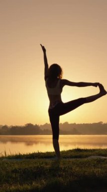 Sahilde yoga yapan, çimenli bir sahilde yaz tatilinin ve güneşin tadını çıkaran, doğadan enerji ve sükunet toplayan genç bir kadının portresi. Yüksek kalite 4k görüntü