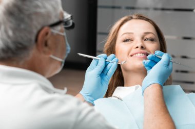 Dişçi koltuğundaki bir hasta, dişçi tedavisiyle yaptığı güzel gülümsemeyi mutlulukla sunar. Prosedürü uygulayan doktor görünüşünden memnun ve bunu sağlamak için çabalıyor.