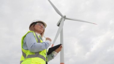 Modern rüzgar türbinlerinin arka planına karşı, dijital tabletli bir kadın enerji santrallerinin üretkenliğini ve sürdürülebilirliğini artırmak için yenilikçi çözümler geliştiriyor. Yüksek kalite 4k görüntü