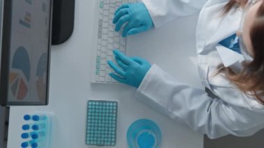 Aşı geliştirme laboratuarında. Teknolojik modern tıbbi analiz laboratuvarı. Beyaz önlüklü bir kadın koruyucu eldivenlerle bilgisayarın başında oturuyor ve yukarıdan klavye görüntüsünü daktilo ediyor. Yüksek