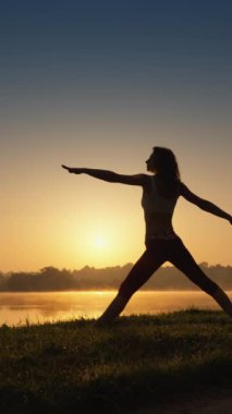 Dikey çekim: Odaklanma ifadesi olan bir kadın gün doğumunda arka planda yoga yapıyor, doğanın büyüsünü hissediyor. Yüksek kalite 4k görüntü