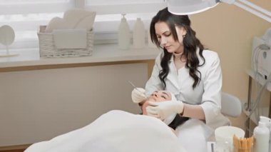 Bir kaplıca kliniğinde profesyonel yüz bakımı yapan deneyimli bir estetisyen. Estetikçi özel aletler ve ürünler kullanırken müşteri bir tedavi yatağında yatıyor..
