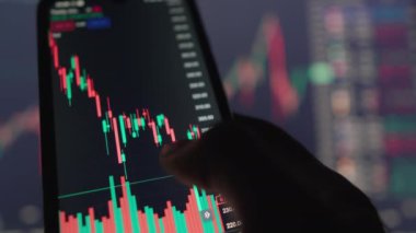 Video çerçeveleri, dinamik borsa verileri ve grafiklerini gösteren bir cep telefonu ekranı ile meşgul olan bir eli betimliyor. Grafiklerde şamdan desenleri ve çeşitli finansal göstergeler yer alıyor.
