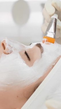 Profesyonel bir yüz tedavisinin yakından görüntüsü, yetenekli bir estetisyen tarafından fırçayla uygulanan beyaz bir maske takan sakin bir müşterinin görüntüsü. Sahnede cilt bakımı ve rahatlığı vurgulanıyor..
