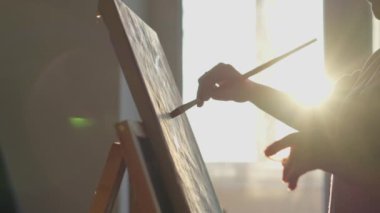 Güneşli bir stüdyoda tuvale resim yapan bir sanatçının ayrıntılı dizisi. Güneş ışığı, fırça darbeleri ve sanatsal odaklanma ile yaratıcı atmosferi güçlendirir..