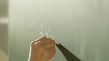 Sınıf ortamında karatahtaya matematiksel denklemler yazan bir öğretmenin yakın çekimi. Sıralama tebeşir yazma hareketini yakalıyor, eğitim ve öğrenme ortamını vurguluyor.