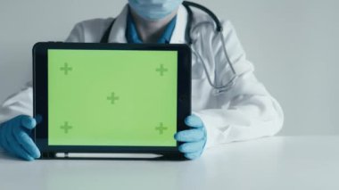 Beyaz önlüklü, maskeli ve eldivenli bir tıp uzmanı elinde çeşitli video çerçevelerinde yeşil ekranlı bir tablet tutuyor. Uygulamalar, sağlık hizmetleri veya teknolojiyle ilgili sunumlar için ideal