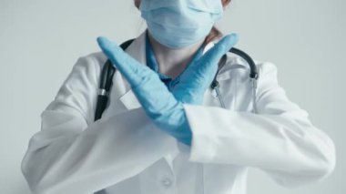 Beyaz önlüklü, mavi eldivenli ve cerrahi maskeli bir kadın doktor, uyarı ya da yasaklama işareti olarak X şeklinde bir dur hareketi yapar. Görüntü ihtiyat ve güvenlik tedbirlerini iletiyor