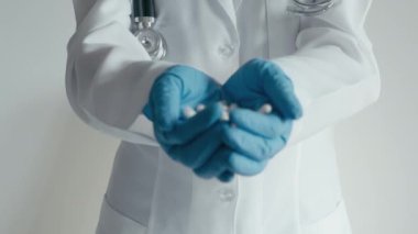 Beyaz laboratuvar önlüklü ve elinde bir avuç dolusu beyaz kapsül tutan tıbbi eldivenli bir doktorun yakından görüntüsü. Bu betimlemede sağlık hizmetleri, profesyonel tıbbi uygulamalar ve ilaç hizmetleri vurgulanıyor.