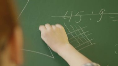 Bir eğitimci, arkasından bakıldığında, yeşil bir karatahtaya matematiksel formüller yazar. Resim dizisi, bir sınıf ortamında öğretim ve matematiksel kavramları açıklama eylemini yakalar..