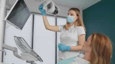 Modern bir diş kliniğinde bir kadın dişçi, diş aletlerini ayarlıyor ve bir hastaya lamba ayarlıyor. Hasta, profesyonel bir sağlık hizmetleri ortamında sağlık kontrolüne hazırdır.