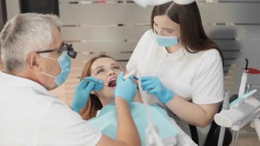 Bir dişçi ve asistanı, modern bir klinikte kadın bir hastaya diş ameliyatı yaparken görülüyor. Maske ve eldiven takıyorlar ve tedavi için steril bir ortam sağlıyorlar..
