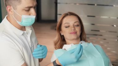 Tıbbi kıyafetli bir dişçi dişçi, dişçi muayenehanesinde kadın bir hastayı tedavi eder. Hasta diş tedavisi gördüğü için rahatlamış görünüyor. Diş hekimi güvenlik için eldiven ve maske takıyor..