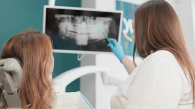 Bir dişçi ve hasta modern bir klinikte diş röntgeni sonuçlarını tartışırken görülüyor. Diş hekimi görüntülerin monitörde görüntülenmesini açıklıyor. Etkili diş bakımı için değerli bilgiler sağlıyor..