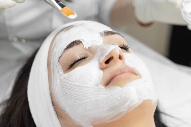 Kaplıca ortamında fırça kullanan bir güzellik uzmanının uyguladığı beyaz krem maskeli profesyonel yüz bakımı alan bir kadının yakın çekimi. Modern bir kaplıcada rahatlama ve cilt bakımı.
