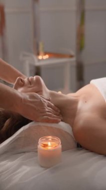 Sakin bir spa ortamında profesyonel boyun masajı terapisinin sakin bir sahnesi. Müşteri beyaz bir havluyla masaj yatağında uzanıyor. Etrafı mum ışığı atmosferiyle çevrili. Rahatlamayı teşvik ediyor..