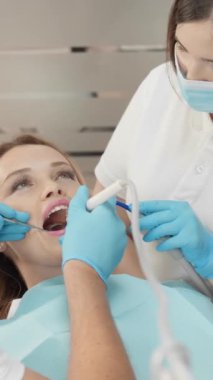 Bir dişçi ve asistanı, modern bir klinikte kadın bir hastaya diş ameliyatı yaparken görülüyor. Maske ve eldiven takıyorlar ve tedavi için steril bir ortam sağlıyorlar..