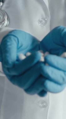 Beyaz laboratuvar önlüklü ve elinde bir avuç dolusu beyaz kapsül tutan tıbbi eldivenli bir doktorun yakından görüntüsü. Bu betimlemede sağlık hizmetleri, profesyonel tıbbi uygulamalar ve ilaç hizmetleri vurgulanıyor.